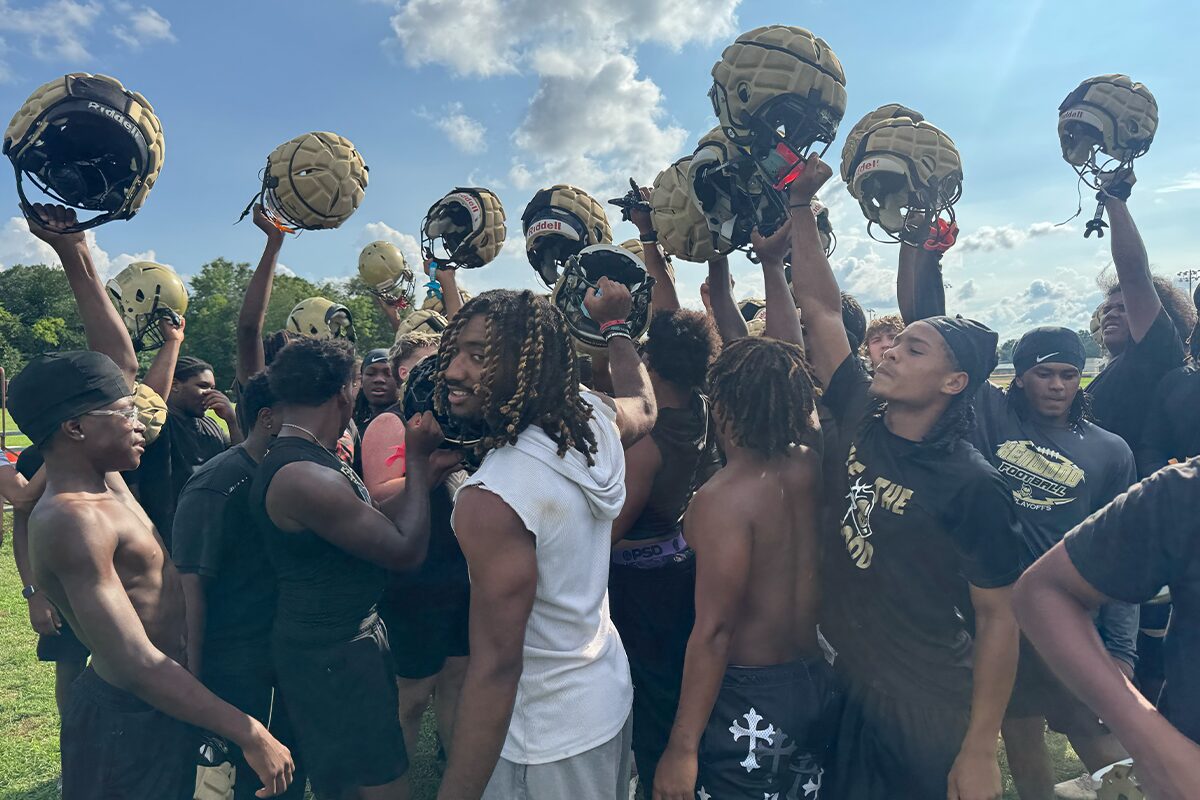 Football players in black uniforms form a huddle, raising gold helmets in celebration on a sunny practice field.