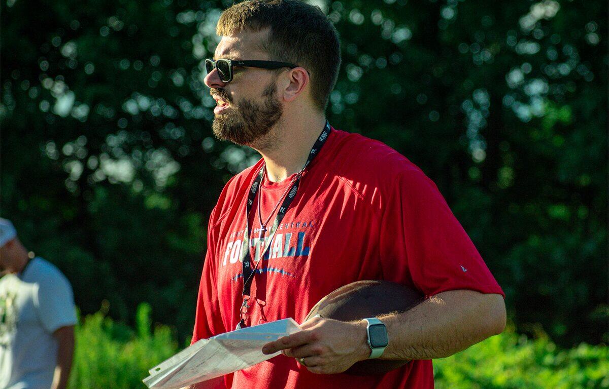 Bearded man in a red shirt and sunglasses holds a football and a clipboard outdoors, with greenery in the background.
