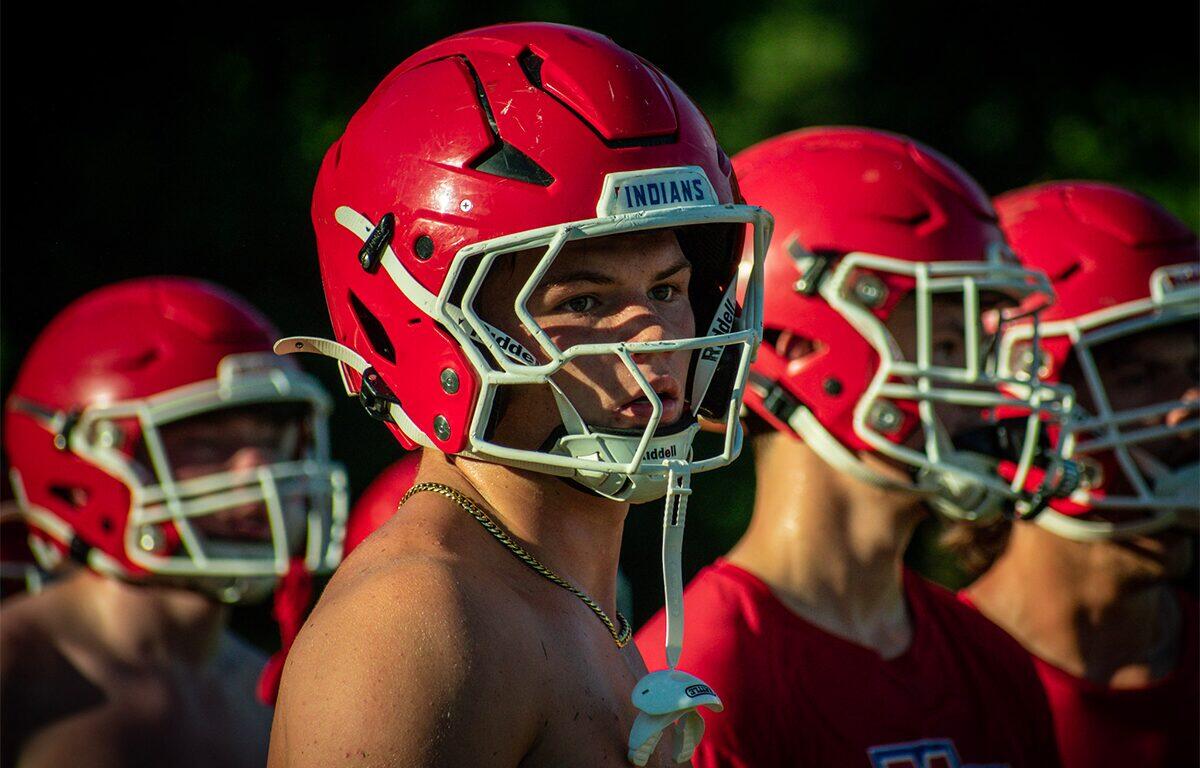 Young football players in red helmets line up during practice, center player facing the camera with a white face mask and chain necklace visible.