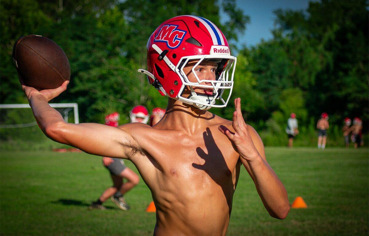Young football quarterback in a red helmet throws a ball on a grassy field during practice.