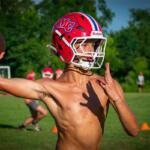 Young football quarterback in a red helmet throws a ball on a grassy field during practice.