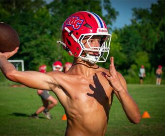 Young football quarterback in a red helmet throws a ball on a grassy field during practice.