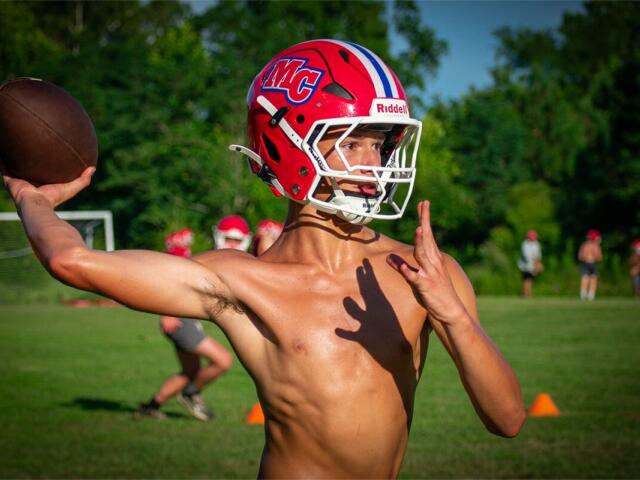 Young football quarterback in a red helmet throws a ball on a grassy field during practice.