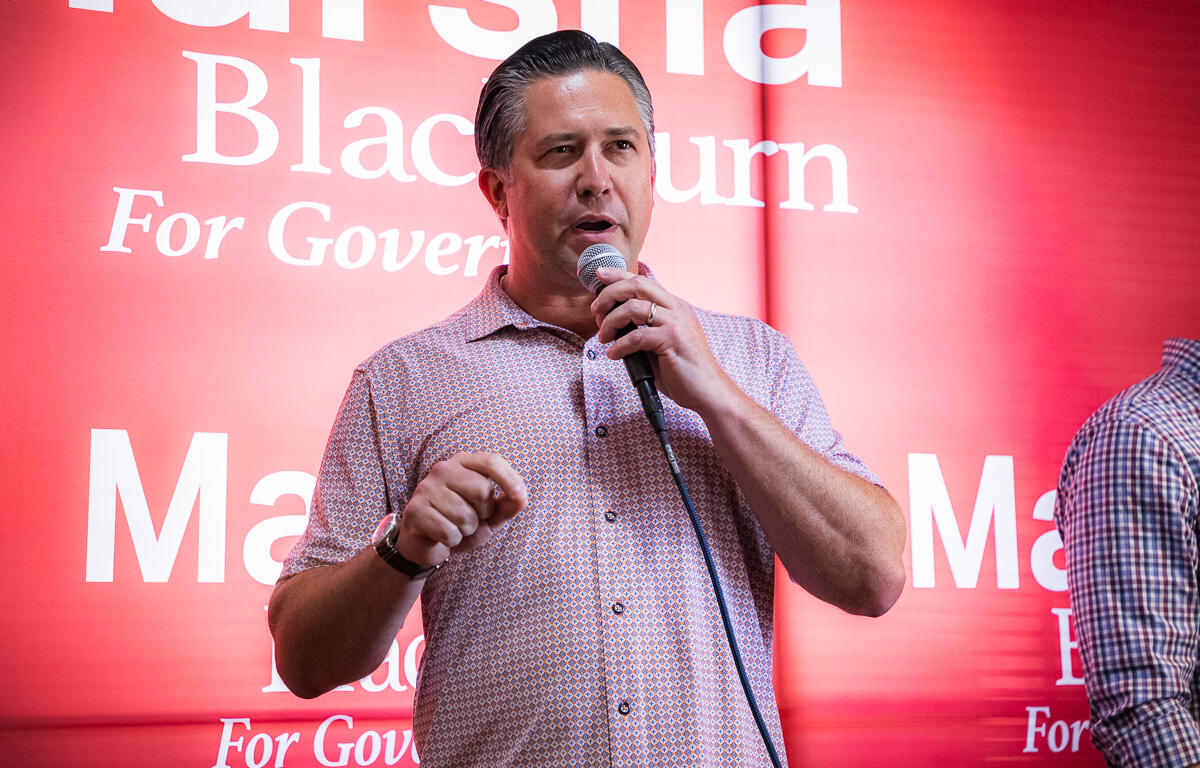 Man speaking into a handheld microphone on stage with a red backdrop and campaign-style text behind him.