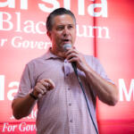 Man speaking into a handheld microphone on stage with a red backdrop and campaign-style text behind him.