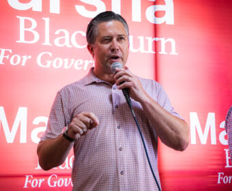 Man speaking into a handheld microphone on stage with a red backdrop and campaign-style text behind him.