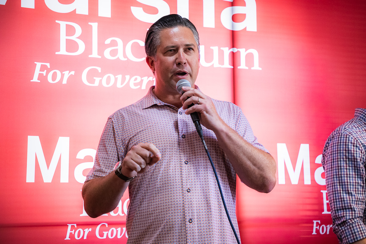 Man speaking into a handheld microphone on stage with a red backdrop and campaign-style text behind him.