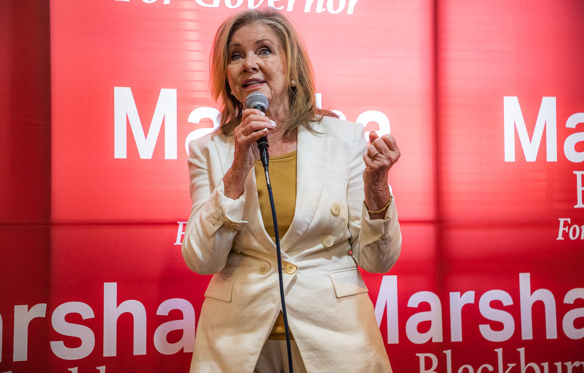 Female political candidate speaking at a podium with a microphone, wearing a light beige blazer over a mustard top, against a red campaign backdrop that reads 'Marshall' and 'For Governor'.