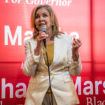 Female political candidate speaking at a podium with a microphone, wearing a light beige blazer over a mustard top, against a red campaign backdrop that reads 'Marshall' and 'For Governor'.