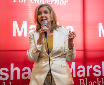 Female political candidate speaking at a podium with a microphone, wearing a light beige blazer over a mustard top, against a red campaign backdrop that reads 'Marshall' and 'For Governor'.