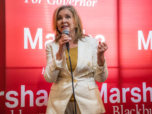 Female political candidate speaking at a podium with a microphone, wearing a light beige blazer over a mustard top, against a red campaign backdrop that reads 'Marshall' and 'For Governor'.
