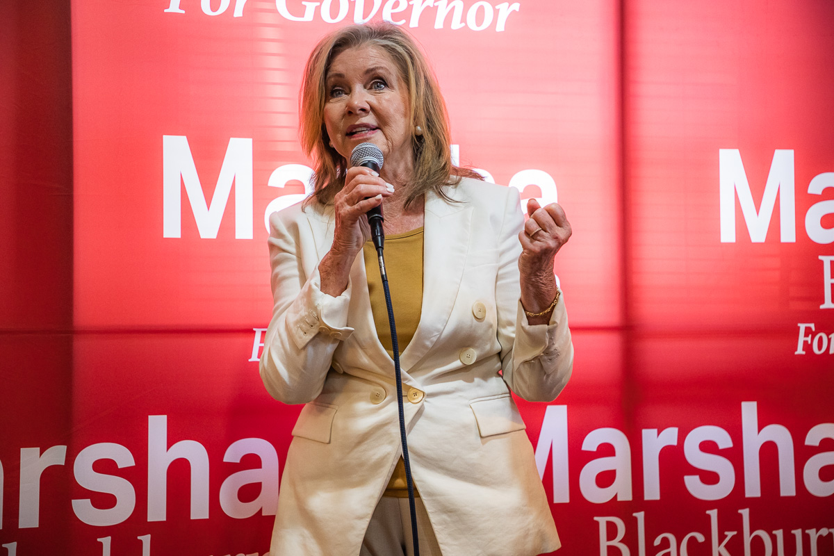 Female political candidate speaking at a podium with a microphone, wearing a light beige blazer over a mustard top, against a red campaign backdrop that reads 'Marshall' and 'For Governor'.