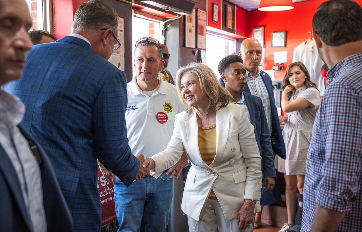 Politician in a white suit shakes hands with a supporter in a blue blazer at a campaign event, surrounded by volunteers in a red-walled room with signs and stickers.