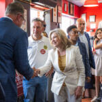 Politician in a white suit shakes hands with a supporter in a blue blazer at a campaign event, surrounded by volunteers in a red-walled room with signs and stickers.
