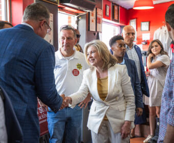 Politician in a white suit shakes hands with a supporter in a blue blazer at a campaign event, surrounded by volunteers in a red-walled room with signs and stickers.