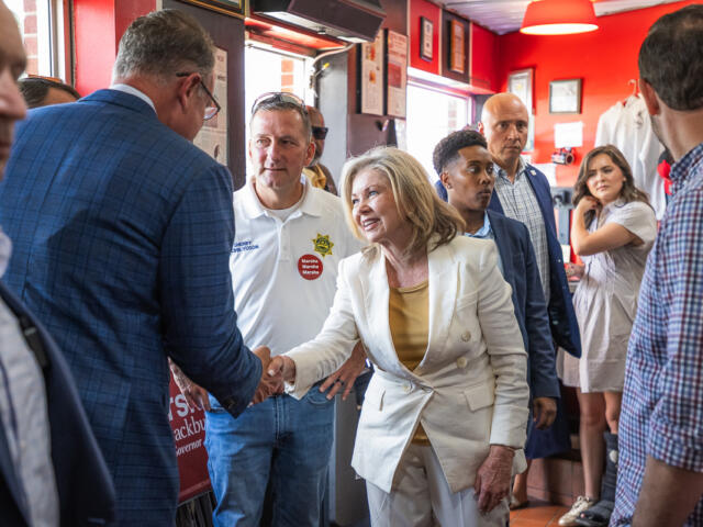 Politician in a white suit shakes hands with a supporter in a blue blazer at a campaign event, surrounded by volunteers in a red-walled room with signs and stickers.