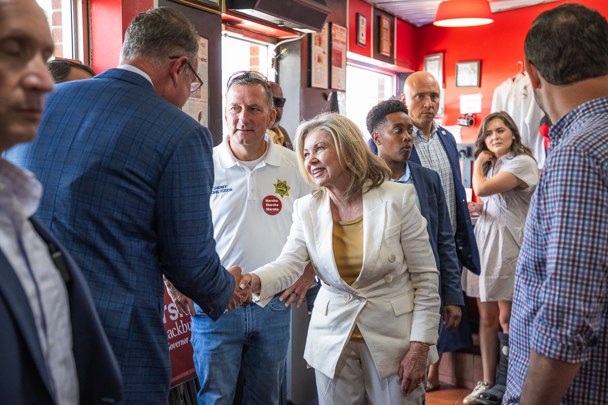 Politician in a white suit shakes hands with a supporter in a blue blazer at a campaign event, surrounded by volunteers in a red-walled room with signs and stickers.