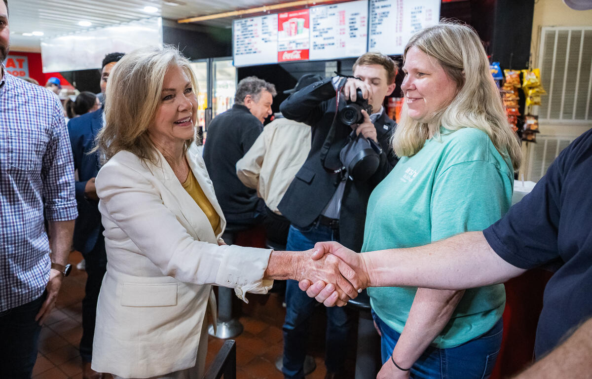 Two women shake hands in a busy cafe or restaurant, while a photographer snaps a picture in the background.