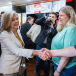 Two women shake hands in a busy cafe or restaurant, while a photographer snaps a picture in the background.