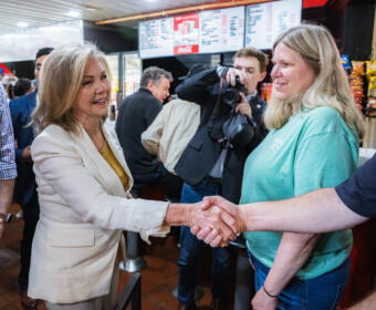 Two women shake hands in a busy cafe or restaurant, while a photographer snaps a picture in the background.