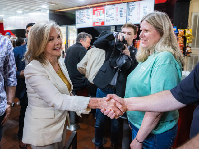 Two women shake hands in a busy cafe or restaurant, while a photographer snaps a picture in the background.