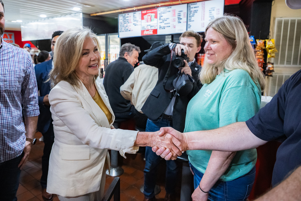 Two women shake hands in a busy cafe or restaurant, while a photographer snaps a picture in the background.