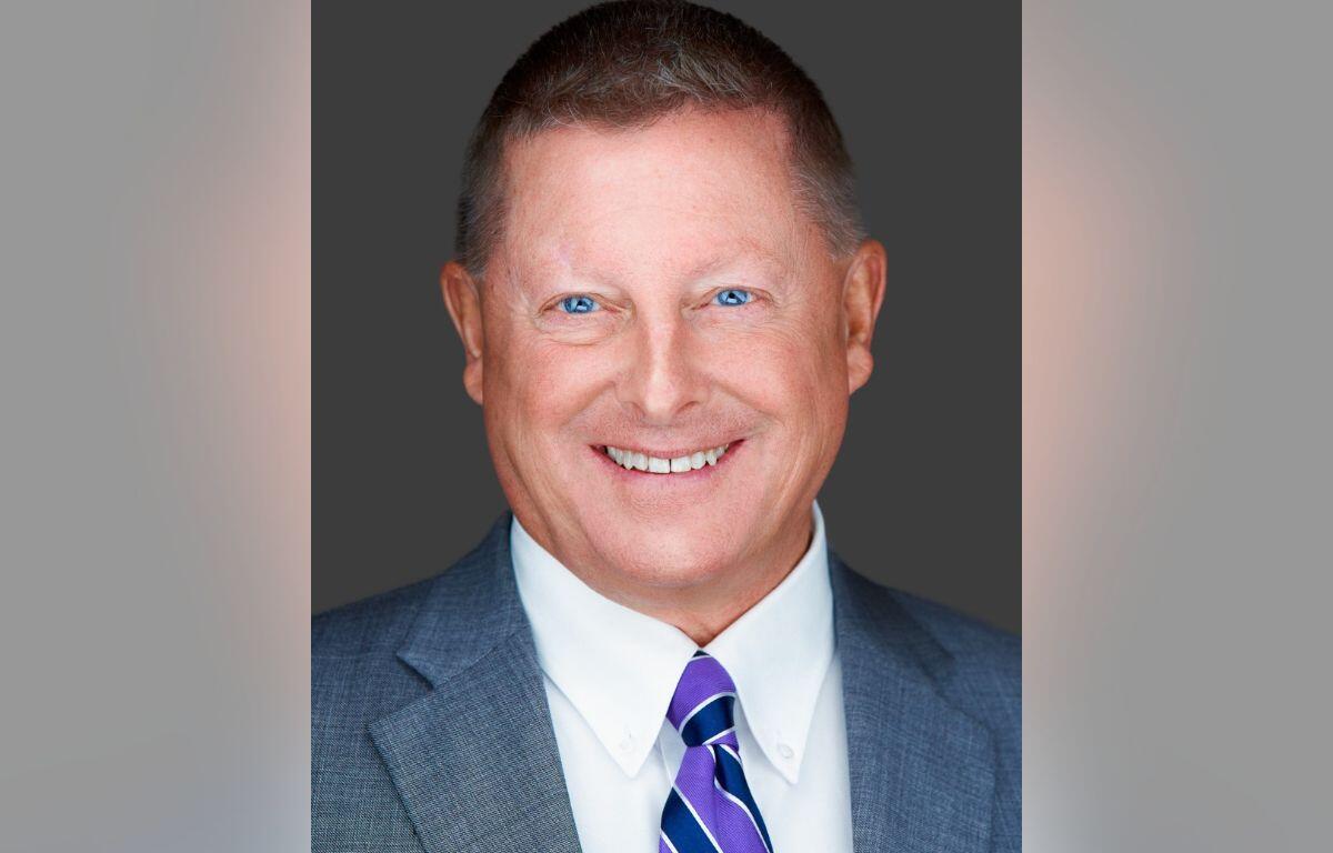 Professional headshot: smiling man in a gray suit, white shirt, and purple-striped tie against a dark backdrop.