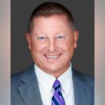 Professional headshot: smiling man in a gray suit, white shirt, and purple-striped tie against a dark backdrop.