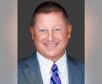 Professional headshot: smiling man in a gray suit, white shirt, and purple-striped tie against a dark backdrop.