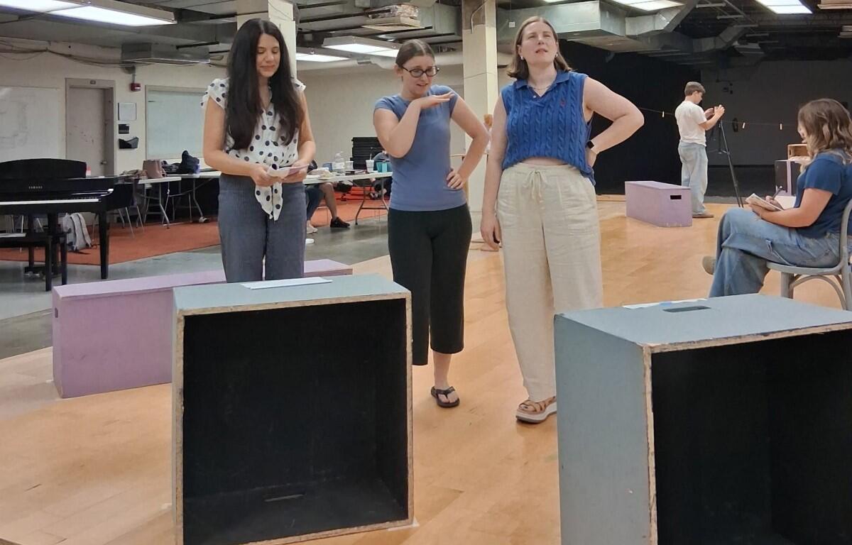 Three women stand and converse in a rehearsal studio, with wooden floor boxes and a piano in the background.