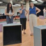 Three women stand and converse in a rehearsal studio, with wooden floor boxes and a piano in the background.