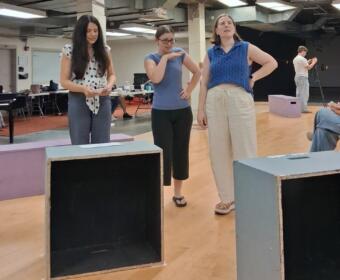 Three women stand and converse in a rehearsal studio, with wooden floor boxes and a piano in the background.