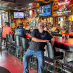 Older man with white beard in a black shirt leaning on a retro bar counter inside a busy, neon-lit tavern; patrons sit at the bar, a dog rests nearby on the left side.