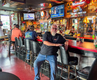 Older man with white beard in a black shirt leaning on a retro bar counter inside a busy, neon-lit tavern; patrons sit at the bar, a dog rests nearby on the left side.