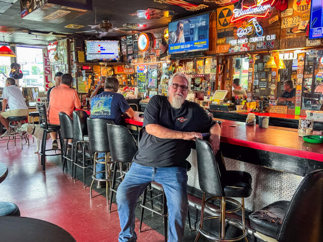 Older man with white beard in a black shirt leaning on a retro bar counter inside a busy, neon-lit tavern; patrons sit at the bar, a dog rests nearby on the left side.