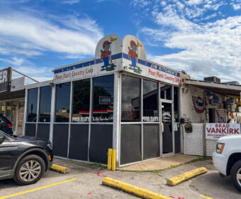 Exterior of Jim Kitchen's Poor Man's Country Club, a small diner with cartoon mascots on the roof and large glass windows in front.