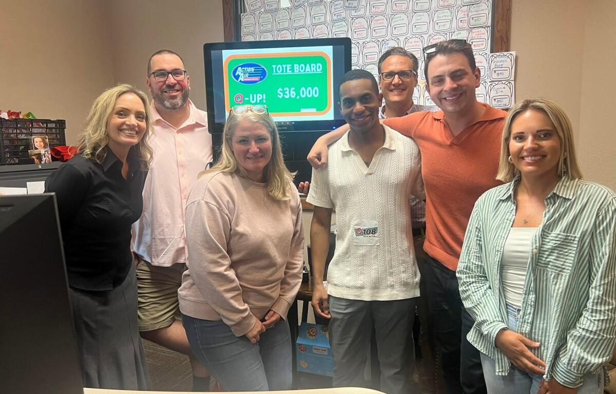 Group of six adults smiling in an office, gathered around a monitor showing a tote board with $36,000 prize.