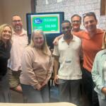 Group of six adults smiling in an office, gathered around a monitor showing a tote board with $36,000 prize.