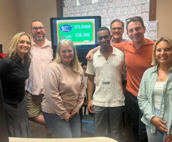 Group of six adults smiling in an office, gathered around a monitor showing a tote board with $36,000 prize.