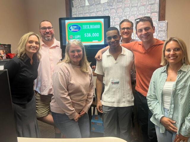 Group of six adults smiling in an office, gathered around a monitor showing a tote board with $36,000 prize.
