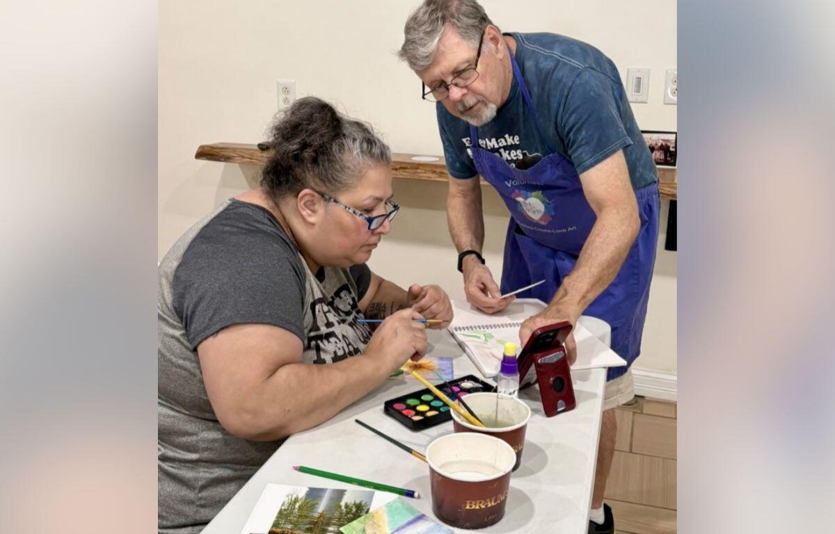 Adult woman seated at a table painting with watercolors while a volunteer in a blue apron assists nearby.