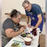 Adult woman seated at a table painting with watercolors while a volunteer in a blue apron assists nearby.