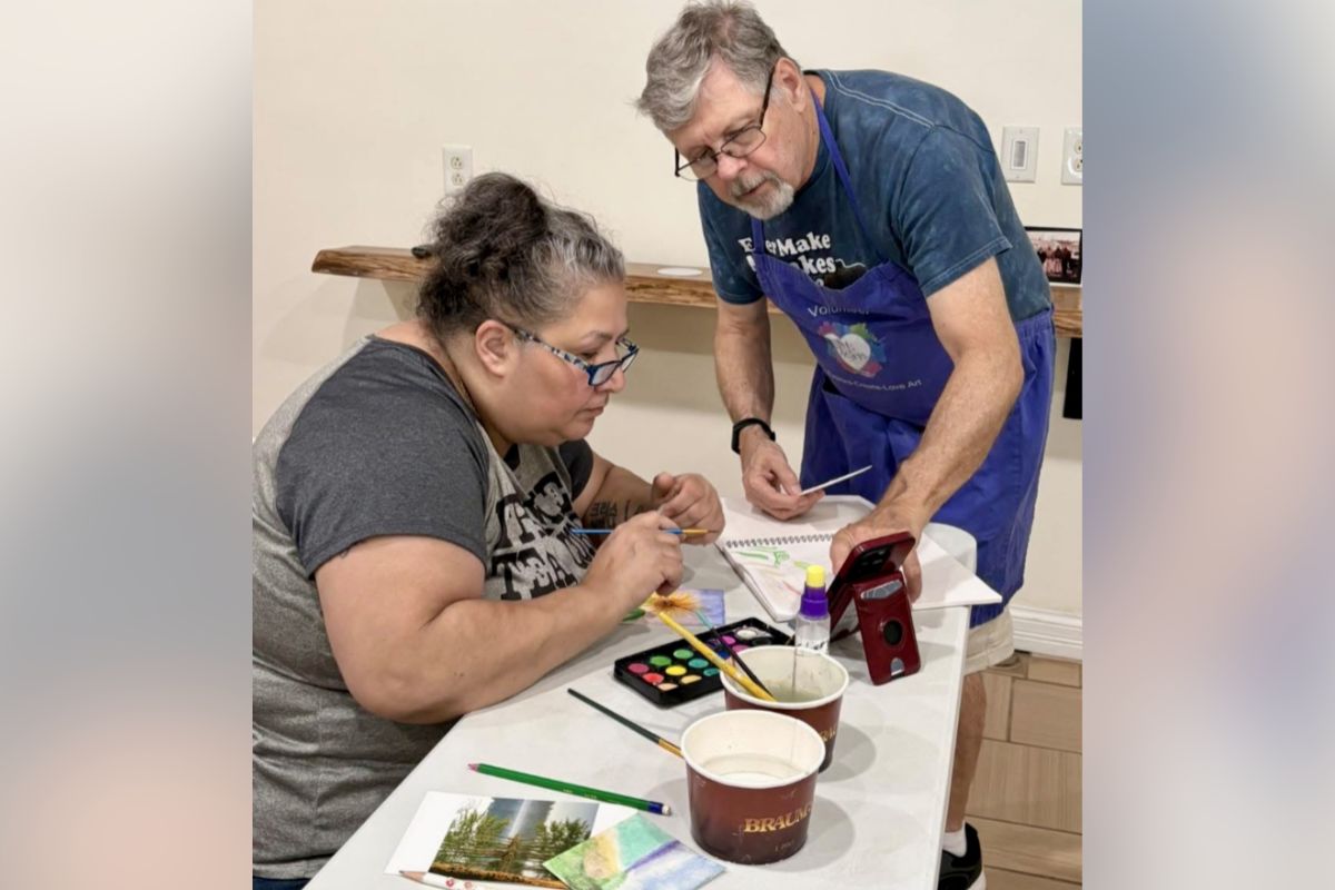Adult woman seated at a table painting with watercolors while a volunteer in a blue apron assists nearby.