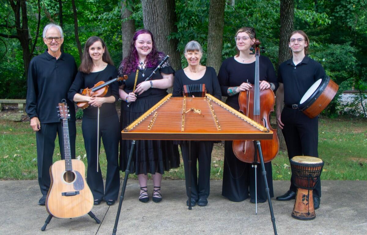 Seven musicians pose outdoors behind a large hammered dulcimer; from left: man with guitar, violinist, woodwind player, center woman, cellist, bassist, and percussionist with a djembe.