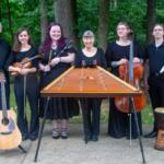 Seven musicians pose outdoors behind a large hammered dulcimer; from left: man with guitar, violinist, woodwind player, center woman, cellist, bassist, and percussionist with a djembe.