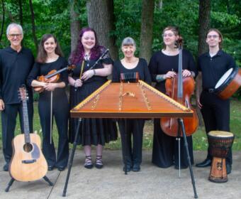 Seven musicians pose outdoors behind a large hammered dulcimer; from left: man with guitar, violinist, woodwind player, center woman, cellist, bassist, and percussionist with a djembe.