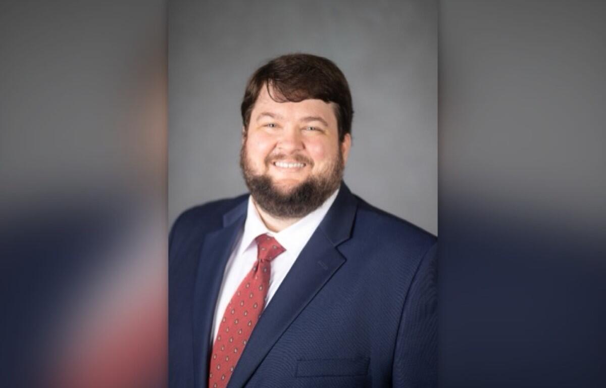 Smiling man in a navy suit and red patterned tie against a gray studio backdrop.