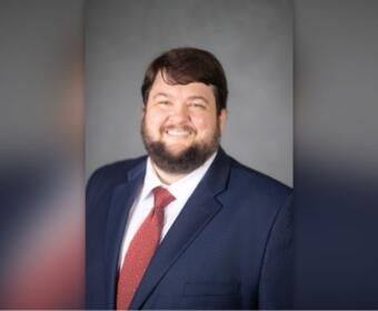 Smiling man in a navy suit and red patterned tie against a gray studio backdrop.