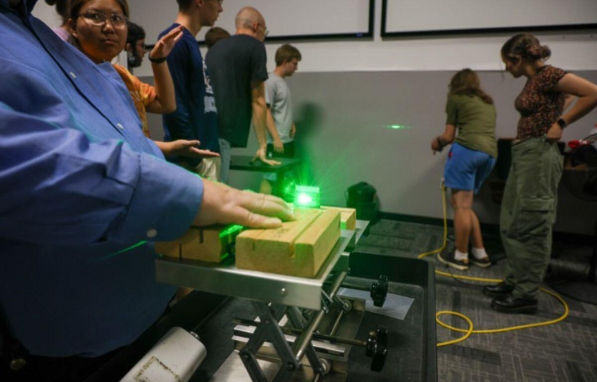 Person in a blue shirt uses a green laser on wooden blocks mounted on a metal track during a classroom demonstration.