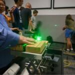 Person in a blue shirt uses a green laser on wooden blocks mounted on a metal track during a classroom demonstration.